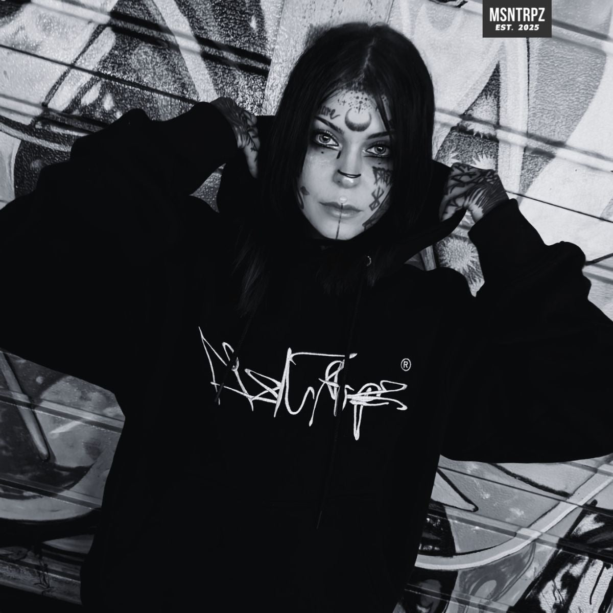 MSNTRPZ® MISANTHROPZ® Noir-Portrait eines Models im schwarzen Oversize-Hoodie vor einer Graffiti-Wand, Kapuze gefasst, markanter Streetwear-Look | Premium German Streetwear