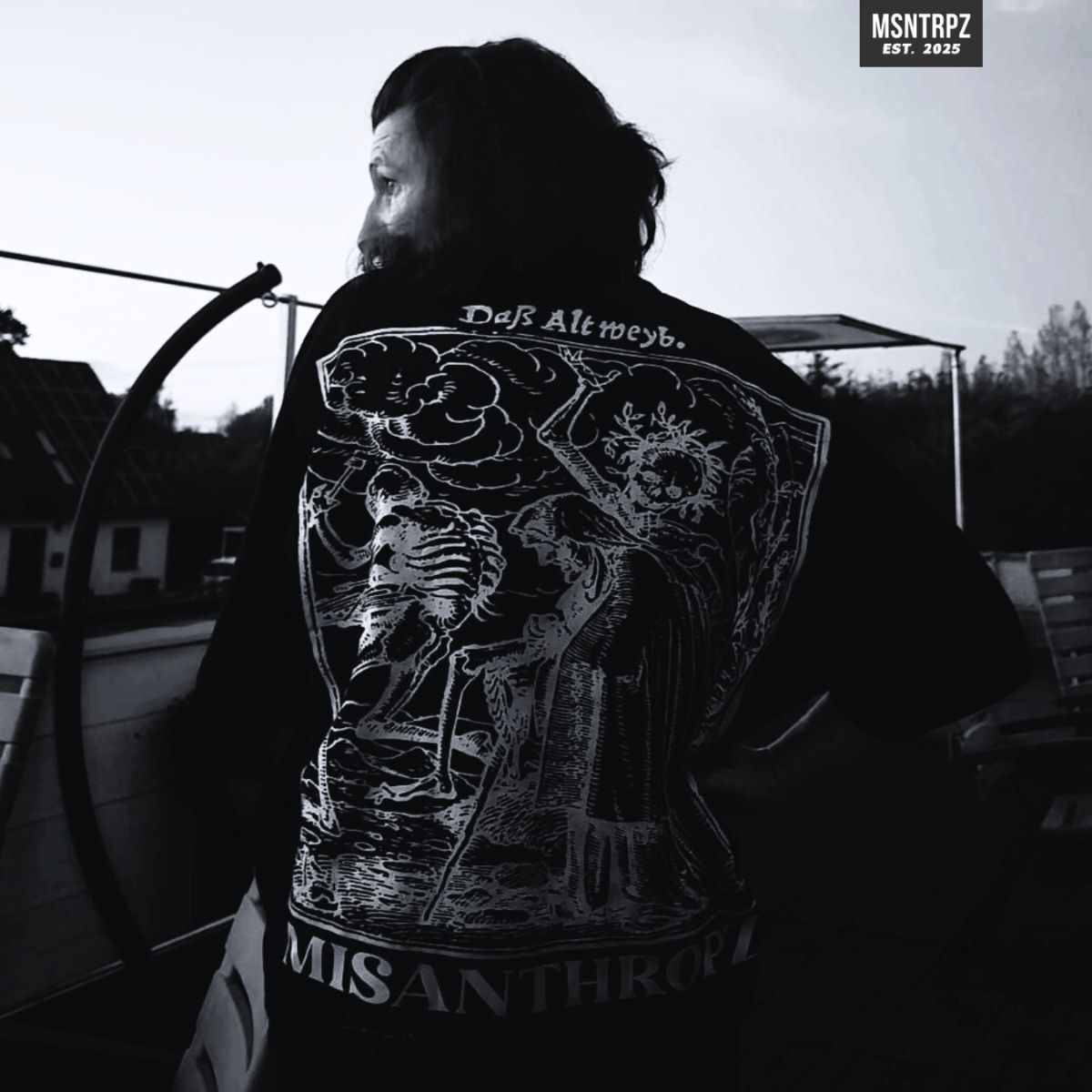 MSNTRPZ® MISANTHROPZ® Noir-Portrait eines langhaarigen Models im Abendlicht, gezeigt von hinten mit dem markanten „Das Altweib.“ Backprint-Shirt. Der ikonische Skelett-Print steht im Kontrast zur ruhigen Outdoorszenerie und verkörpert die rohe, outsider-orientierte Ästhetik der Misanthropz® Bewegung – wenige Souls, echte Tiefe, Premium German Streetwear.