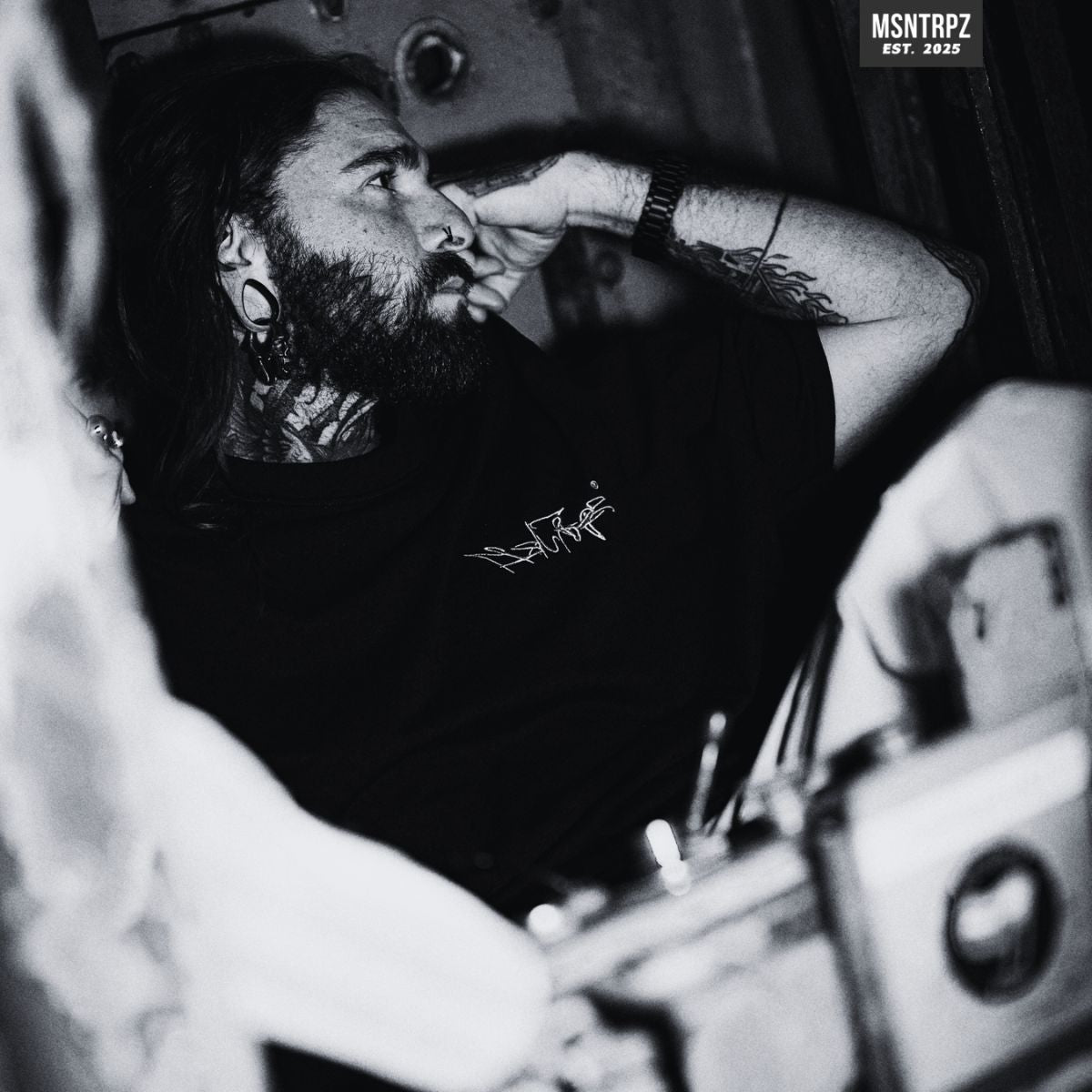 MSNTRPZ® MISANTHROPZ® Noir-Porträt eines tätowierten Models in entspannter Pose, getragenes schwarzes Premium Streetwear-Shirt mit Signature-Frontlogo | Premium German Streetwear