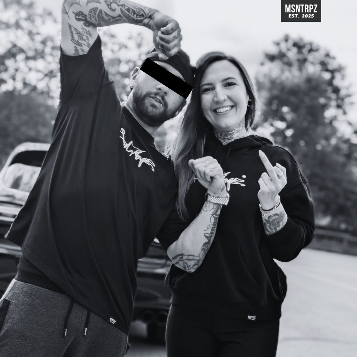 Zwei Models tragen MSNTRPZ® MISANTHROPZ® Streetwear-Hoodies in einer Noir-Outdoor-Aufnahme; lockere Pose vor Fahrzeugkulisse | Premium German Streetwear