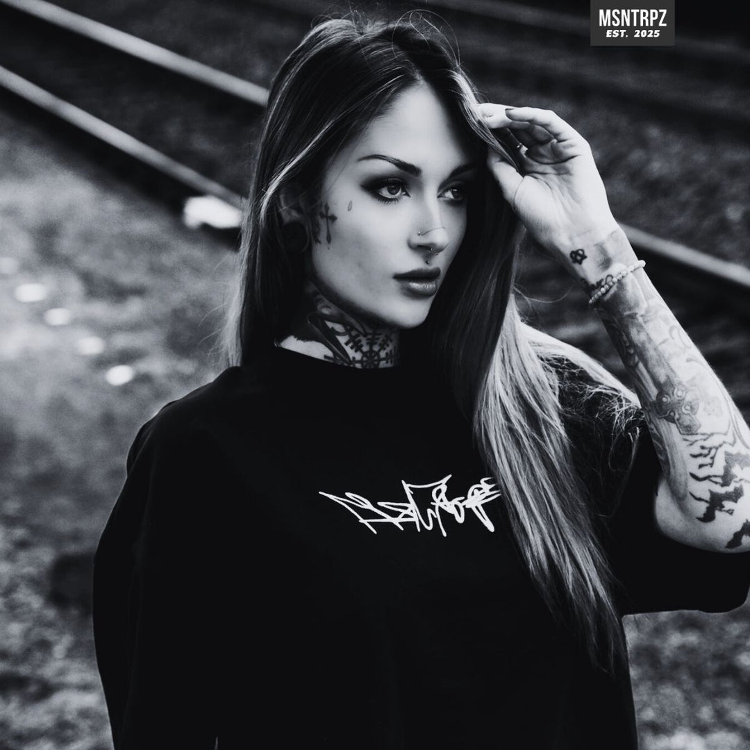 MSNTRPZ® MISANTHROPZ® Noir-Portrait einer tätowierten jungen Frau im schwarzen Premium-Streetwear-Shirt an Bahngleisen – Premium German Streetwear in charakteristischem Dark-Aesthetic-Look.