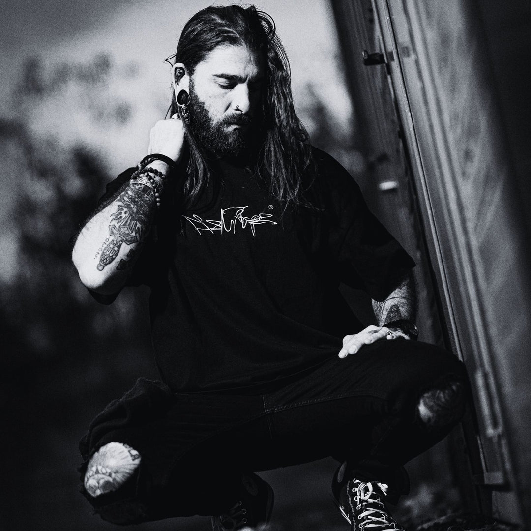 MSNTRPZ® MISANTHROPZ® Tattoo-Model im schwarzen Premium-Streetwear-Shirt, kraftvolle Noir-Aufnahme im knienden Pose – Premium German Streetwear mit authentischer Underground-Energie.