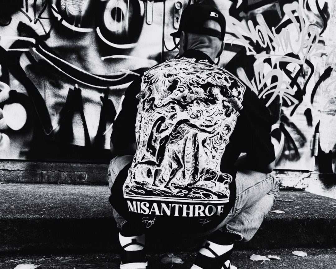 Schwarz-weißes Streetwear-Foto: Person mit MSNTRPZ Premium-Shirt kniet vor einer Graffiti-Wand. Auf dem Rücken ist das ikonische ‚Vertreibung aus dem Paradies‘-Motiv im Barockstil zu sehen – Symbol für Rebellion, Schuld und Erlösung in der MSNTRPZ Kollektion.